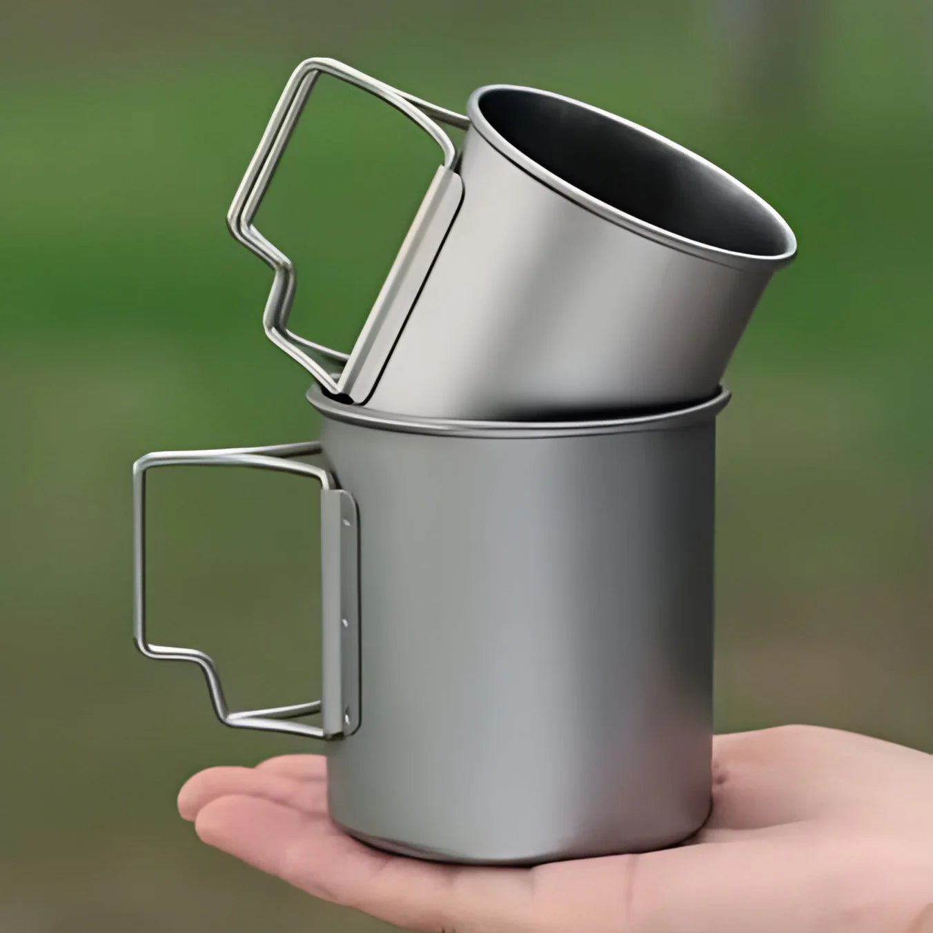 Two metal camping mugs stacked on a hand with a blurred green background