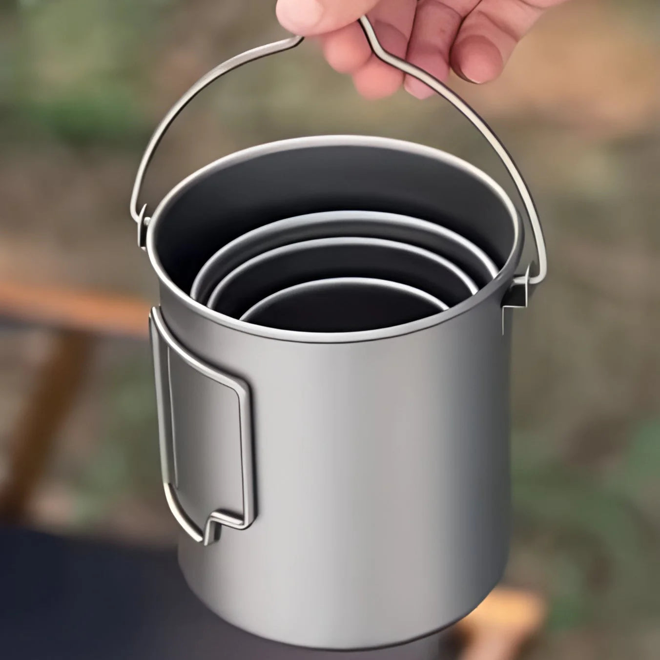 Metal camping pot with handles held by a hand against a blurred natural background
